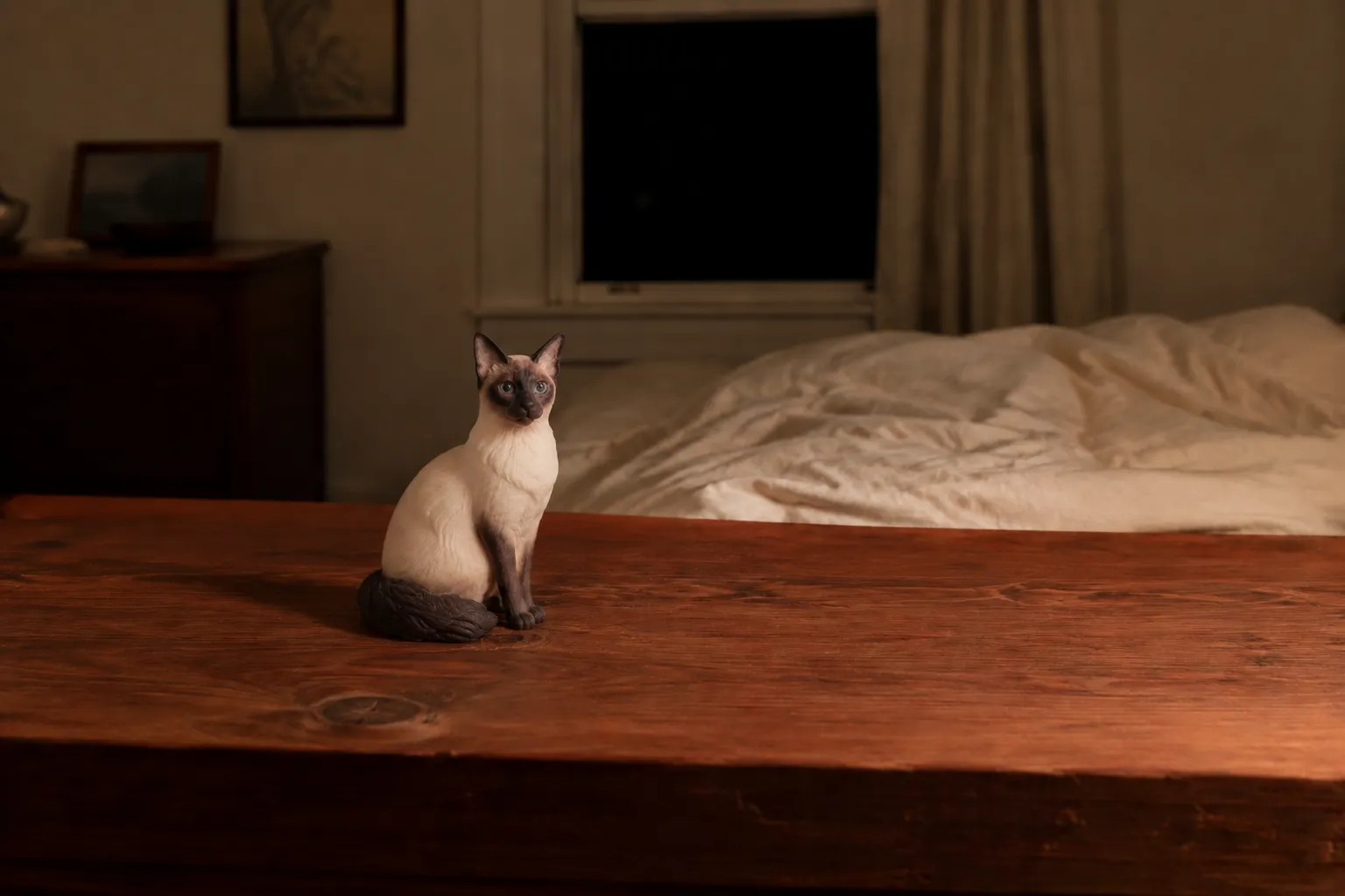 A small Siamese cat figurine, curled asleep, on a cedar chest at the foot of a bed beside a collar and folded blanket.