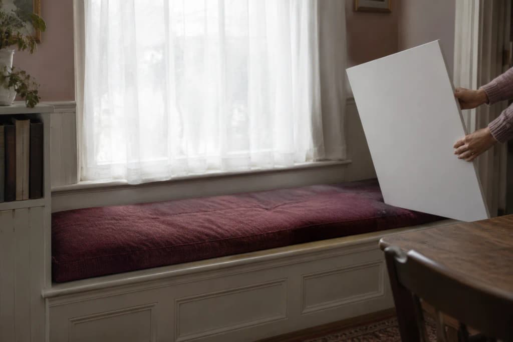 Hands hold a white foam board beside an empty tweed dog cushion on a window seat under a voile-covered window